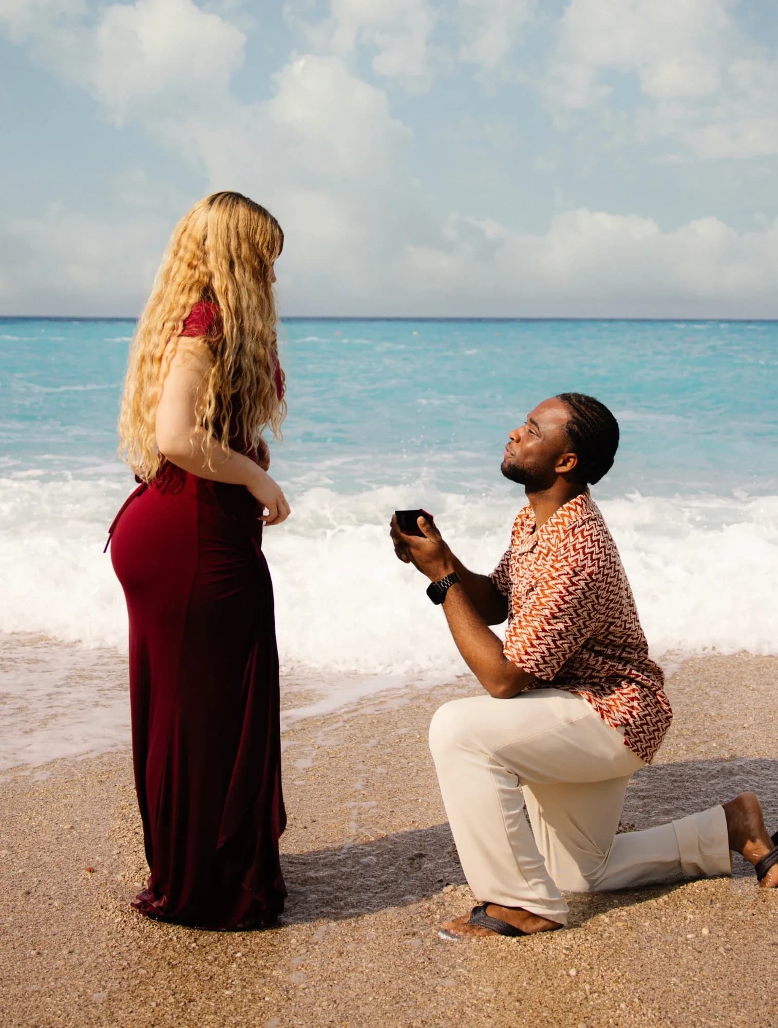 Ese on one knee on a beach, holding out a ring box to Alicia, with the sea behind them